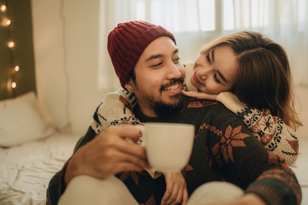 A bearded man in a winter sweater is holding a cup of coffee while being hugged from behind by a woman in a winter sweater.