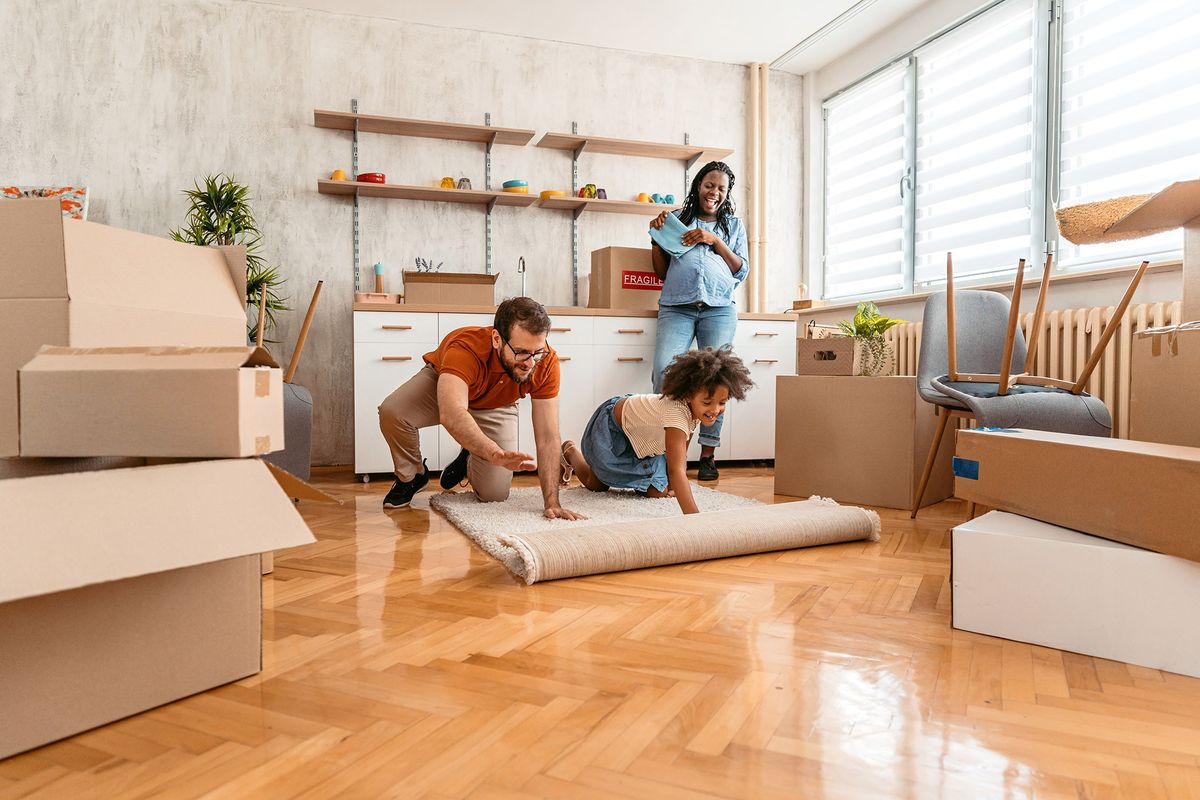 A dad and child roll out a carpet over a clean wooden floor while a pregnant mom watches.