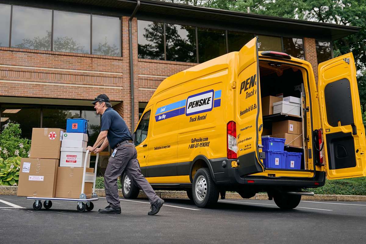 A delivery driver pushes a cart of boxes from a yellow cargo van into a business building.