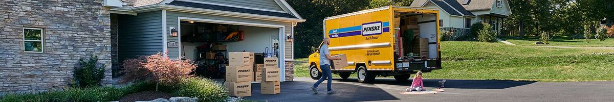 A family loads Penske moving boxes into a yellow Penske truck parked in their driveway.