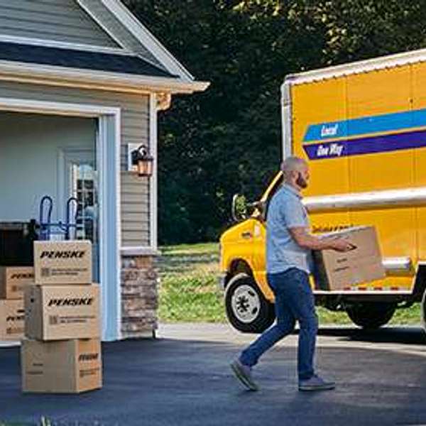 A family loads Penske moving boxes into a yellow Penske truck parked in their driveway.