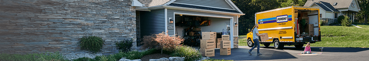 A family loads Penske moving boxes into a yellow Penske truck parked in their driveway.