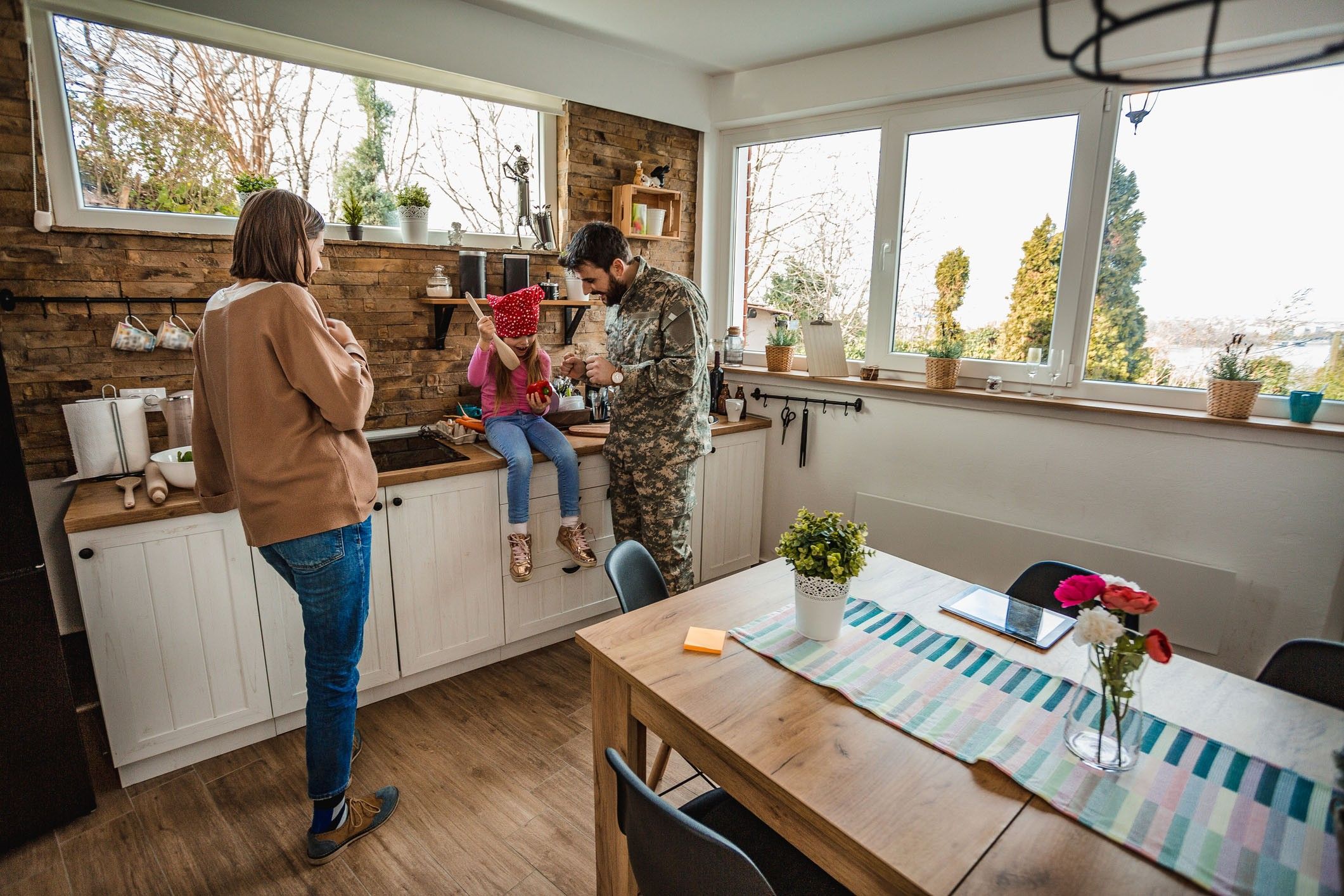 A family of three, one in army fatigues, stands in a cozy kitchen surrounded by windows.
