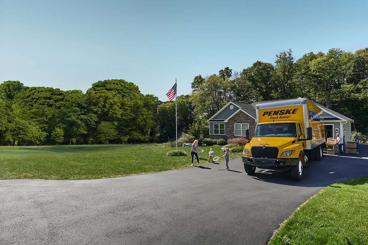 A family with two kids play in the driveway of a house next to a yellow Penske truck.