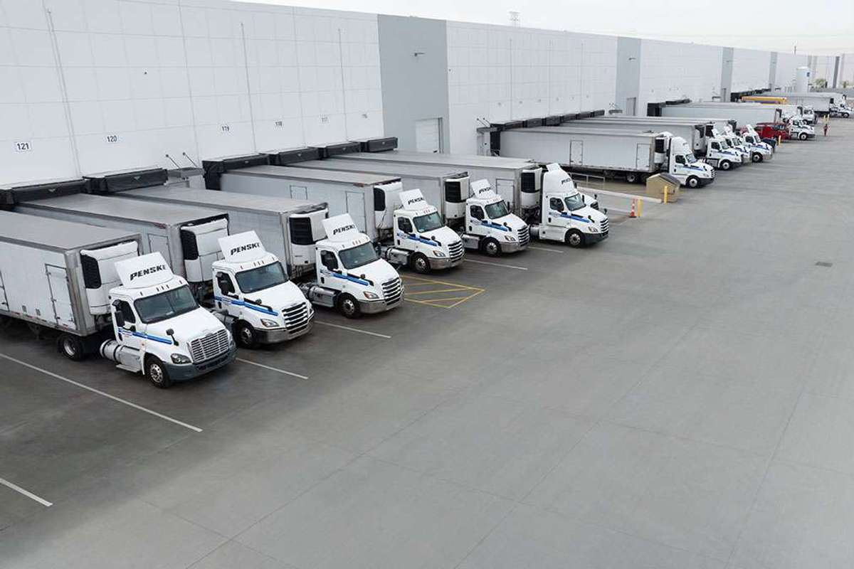 A fleet of white Penske semi-trucks are parked in a line backed up to a big warehouse loading dock.