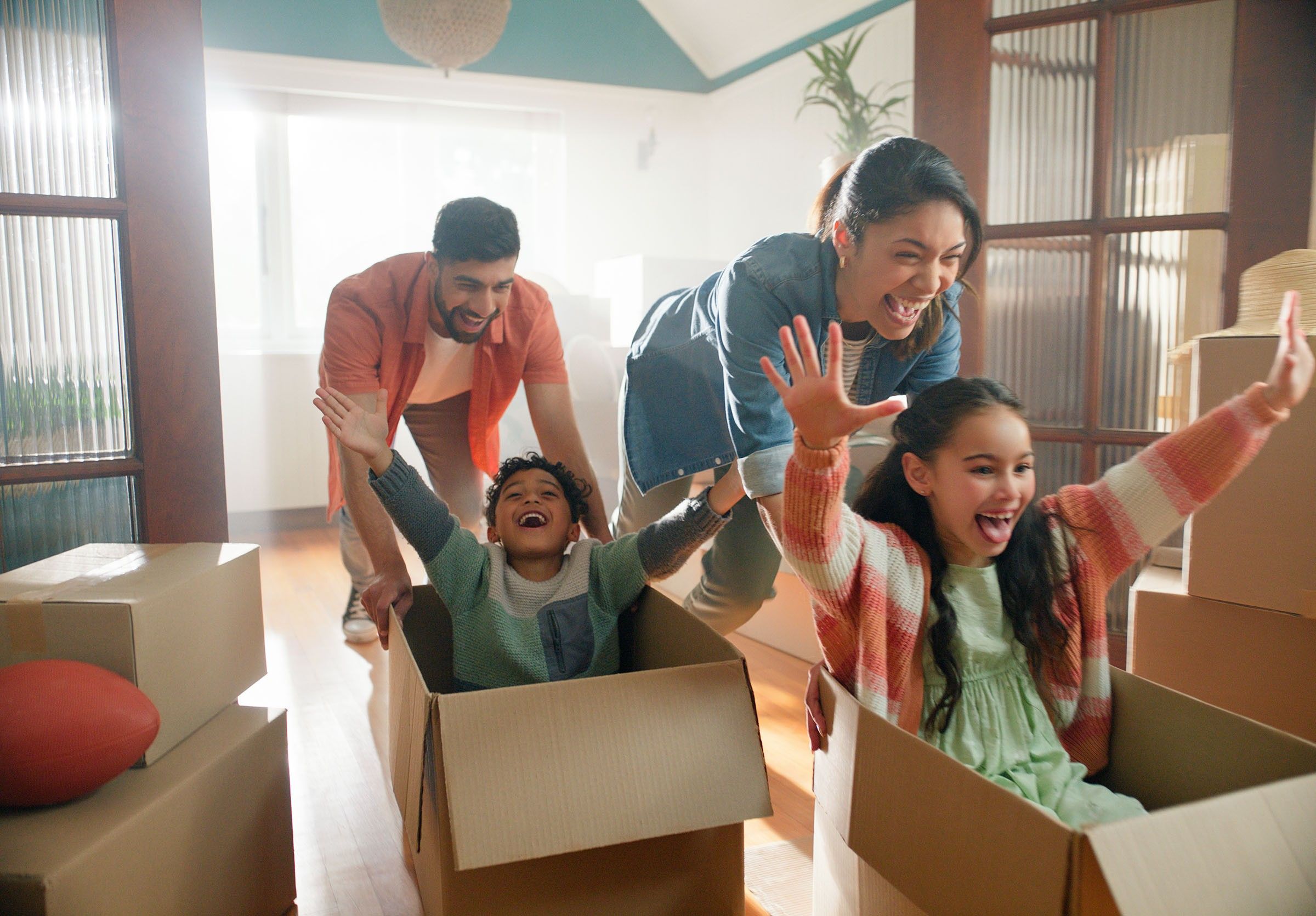 A girl and boy are sitting in empty moving boxes, being pushed around the house by their parents.