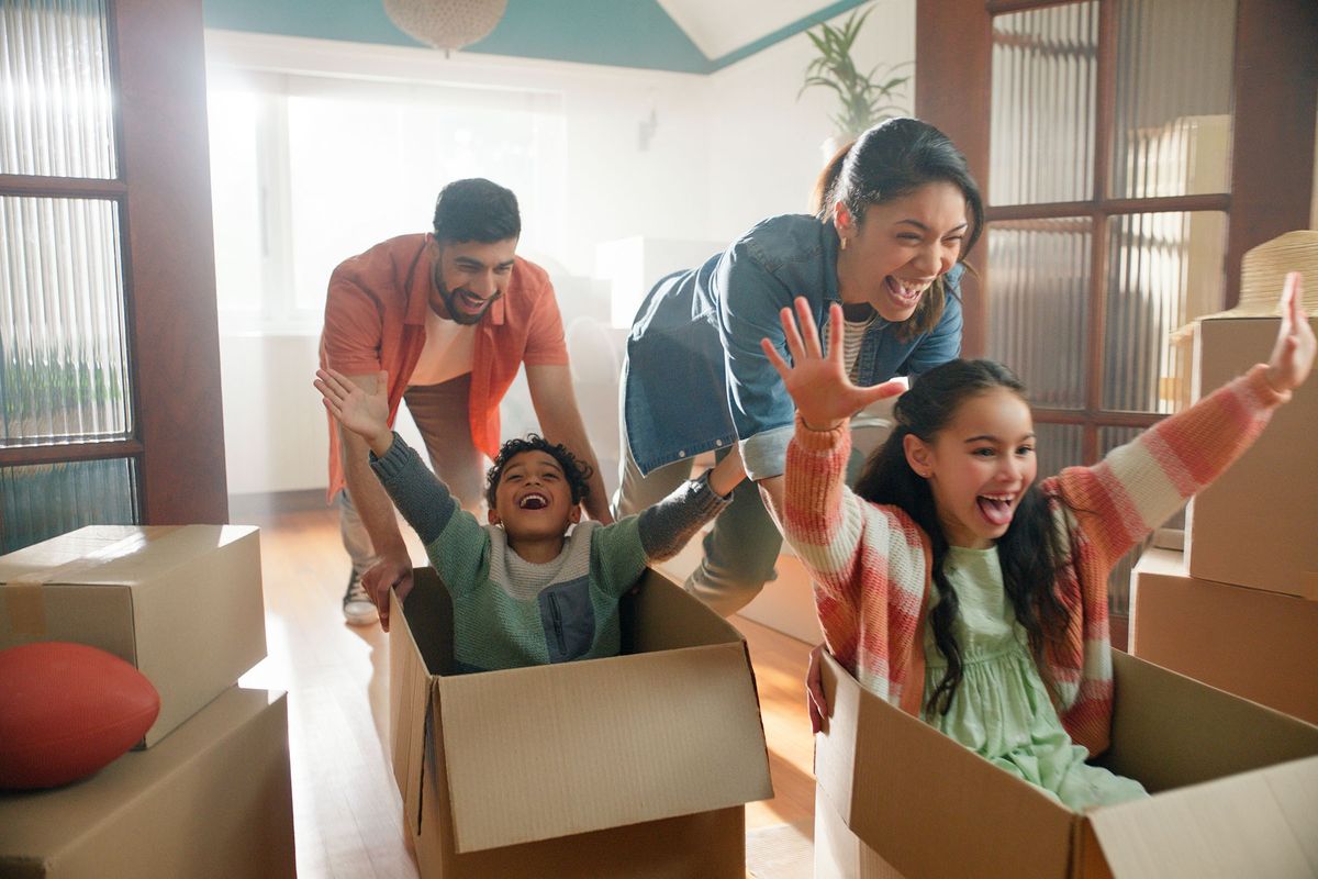 A girl and boy are sitting in empty moving boxes, being pushed around the house by their parents.