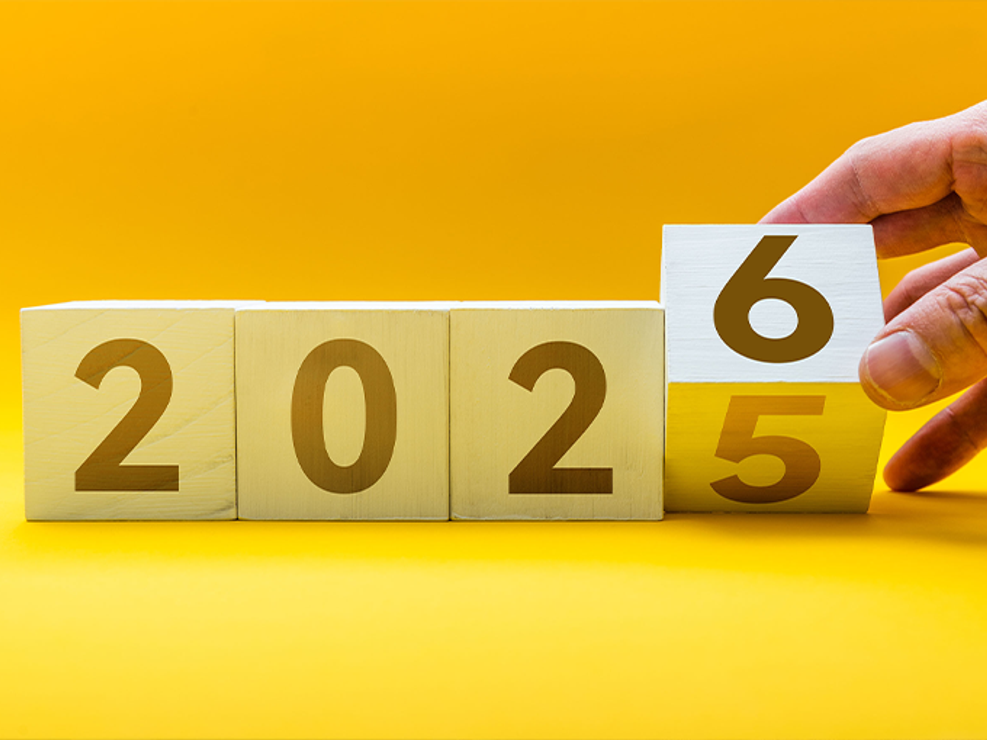 A hand moving blocks from 2025 to 2026 on a yellow background.