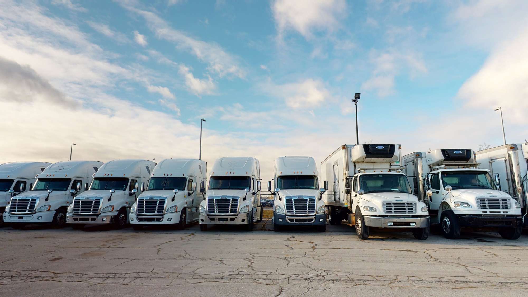 A line of white semi-trucks and box trucks parked in a lot against a blue cloudy sky.