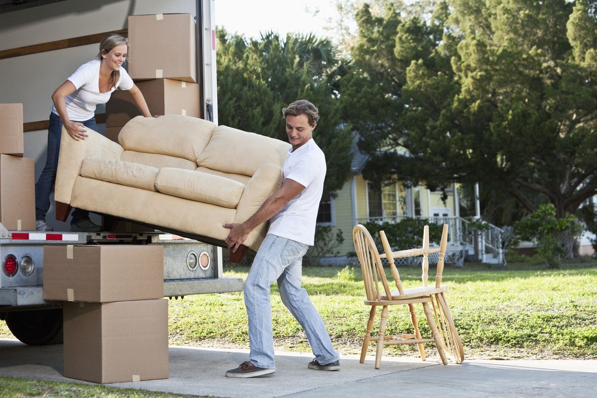 A man and woman carry a white loveseat off of the back of a moving truck.