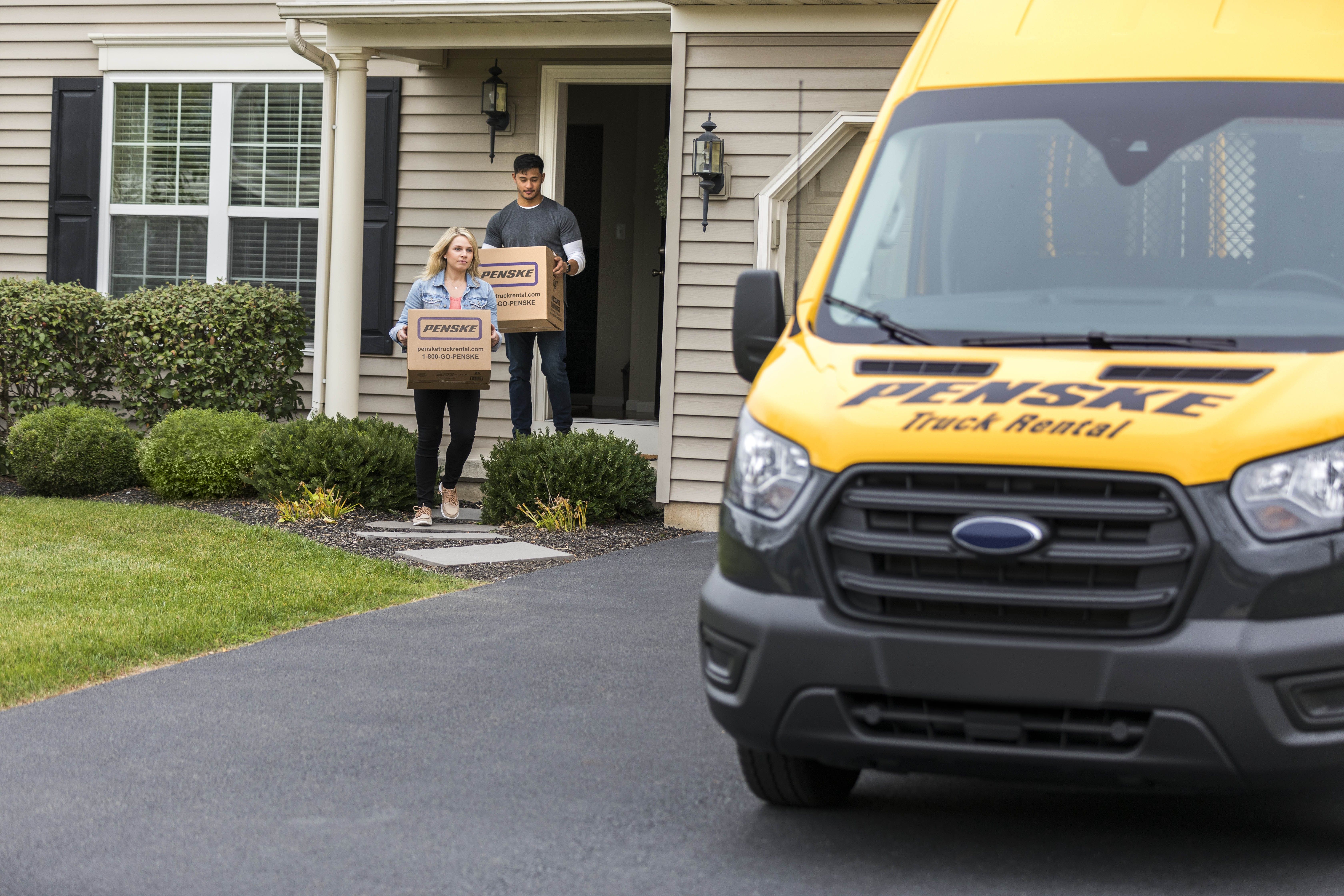 A man and woman carry Penske boxes out of the house to a yellow moving van.