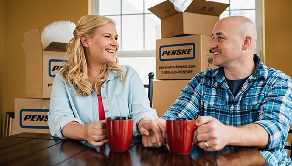 A man and woman sit at a table holding red coffee mugs with Penske moving boxes in the background.