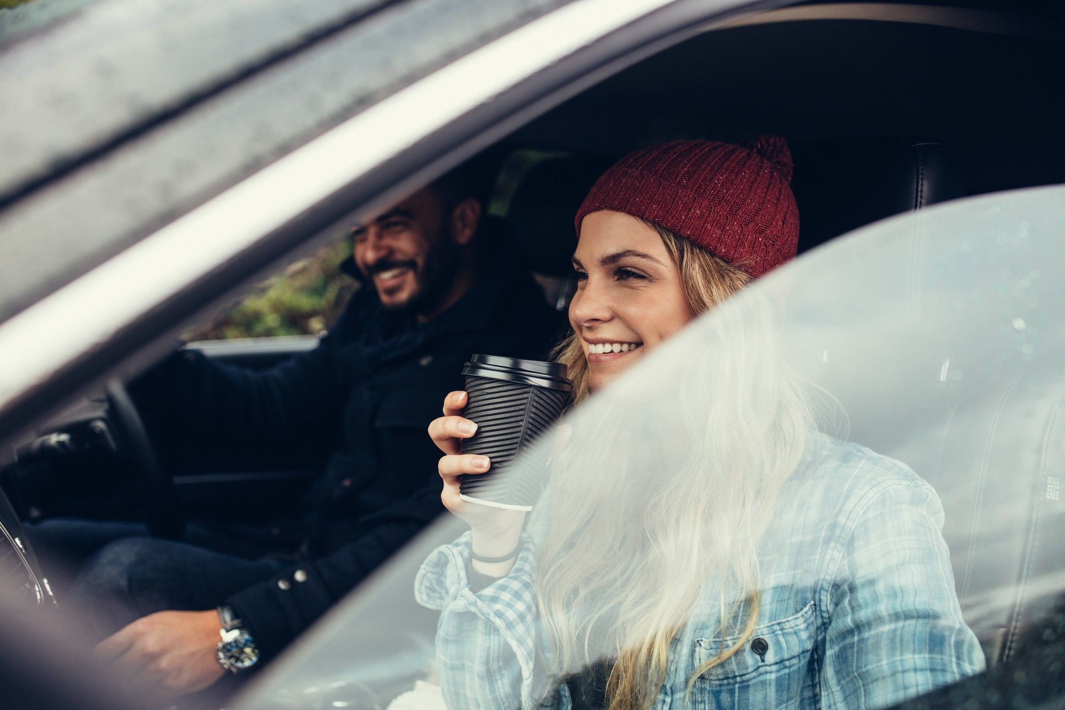 A man and woman sit in the front seat of a rental van, drinking coffee.