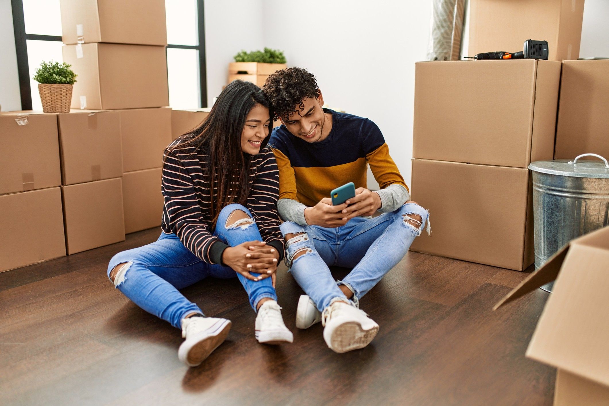 A man and woman sit on the floor amid moving boxes looking at a phone.