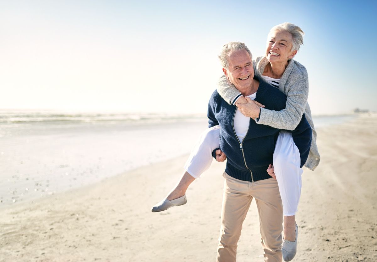 A man carrying an older woman on his back, laughing on the beach.