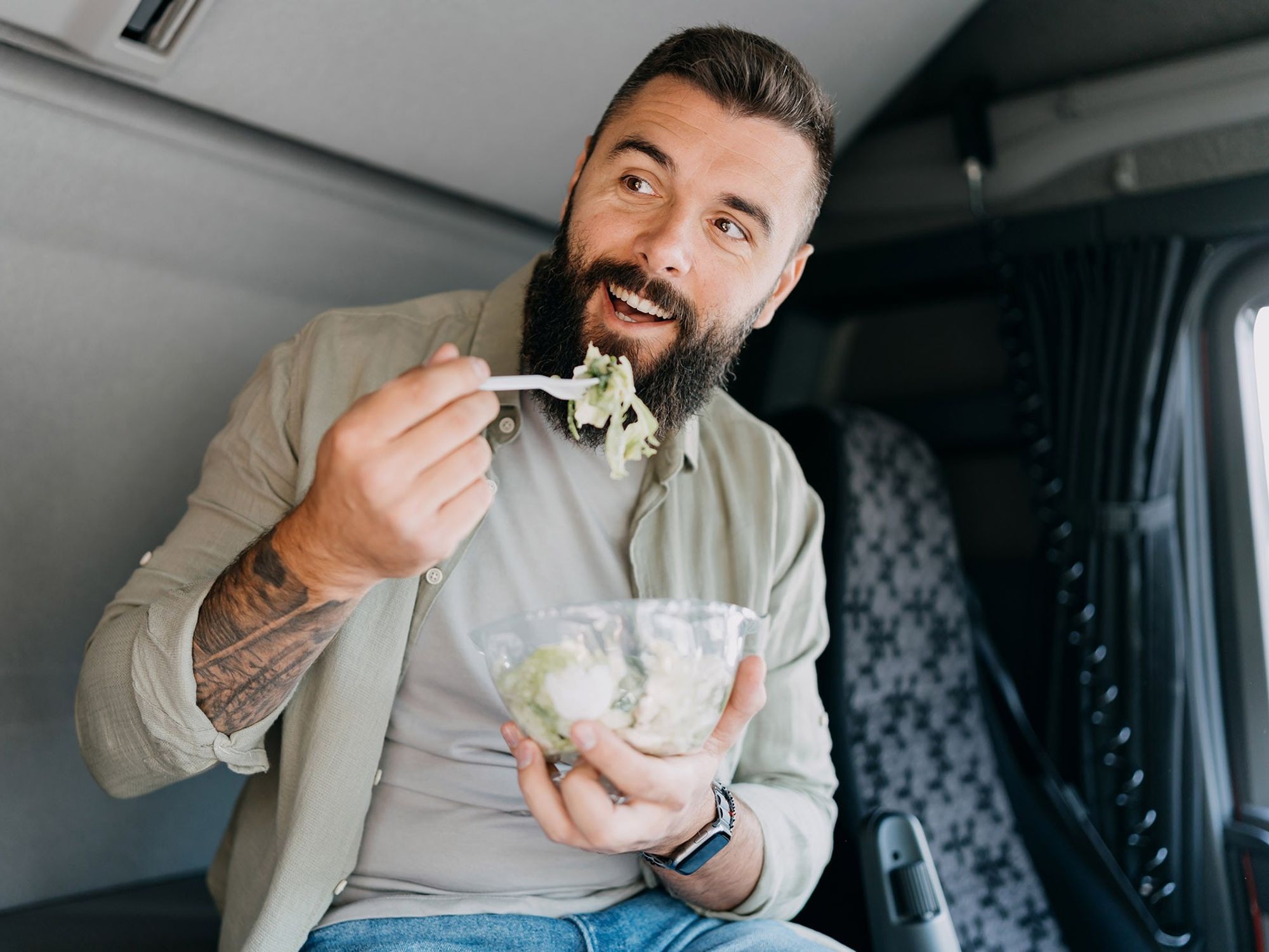 A man eats a salad while sitting in the cab of a semi-truck.