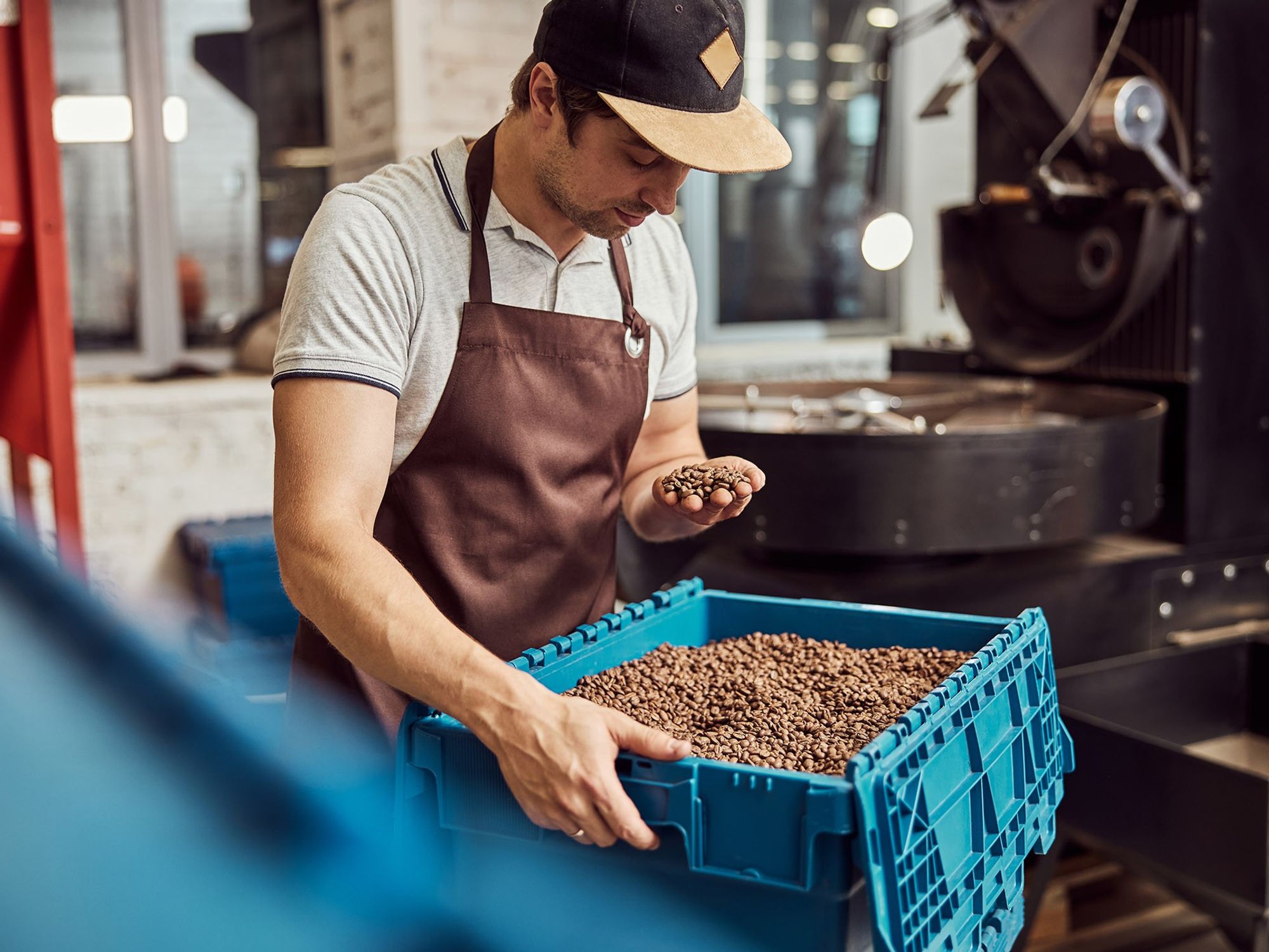 A man holds coffee beans in one hand and a blue bin of coffee beans in the other.
