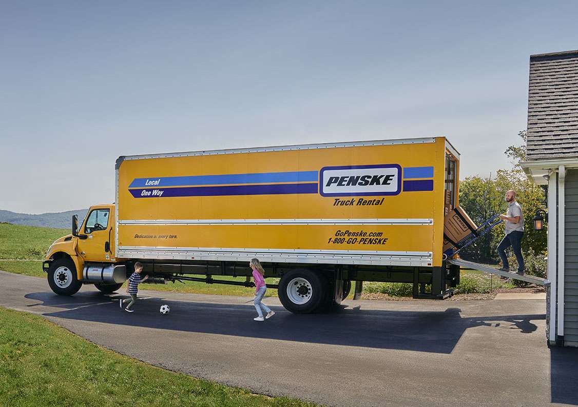 A man pushes boxes on a hand truck up the ramp of a yellow Penske box truck.