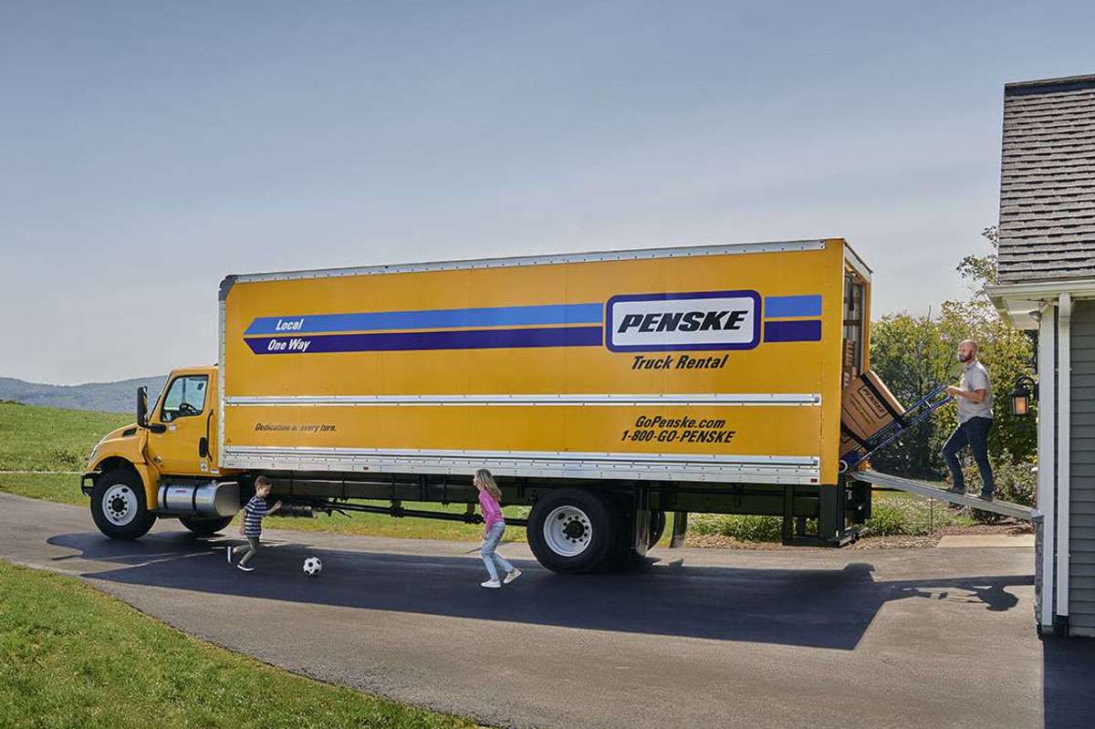 A man pushes boxes on a hand truck up the ramp of a yellow Penske box truck.