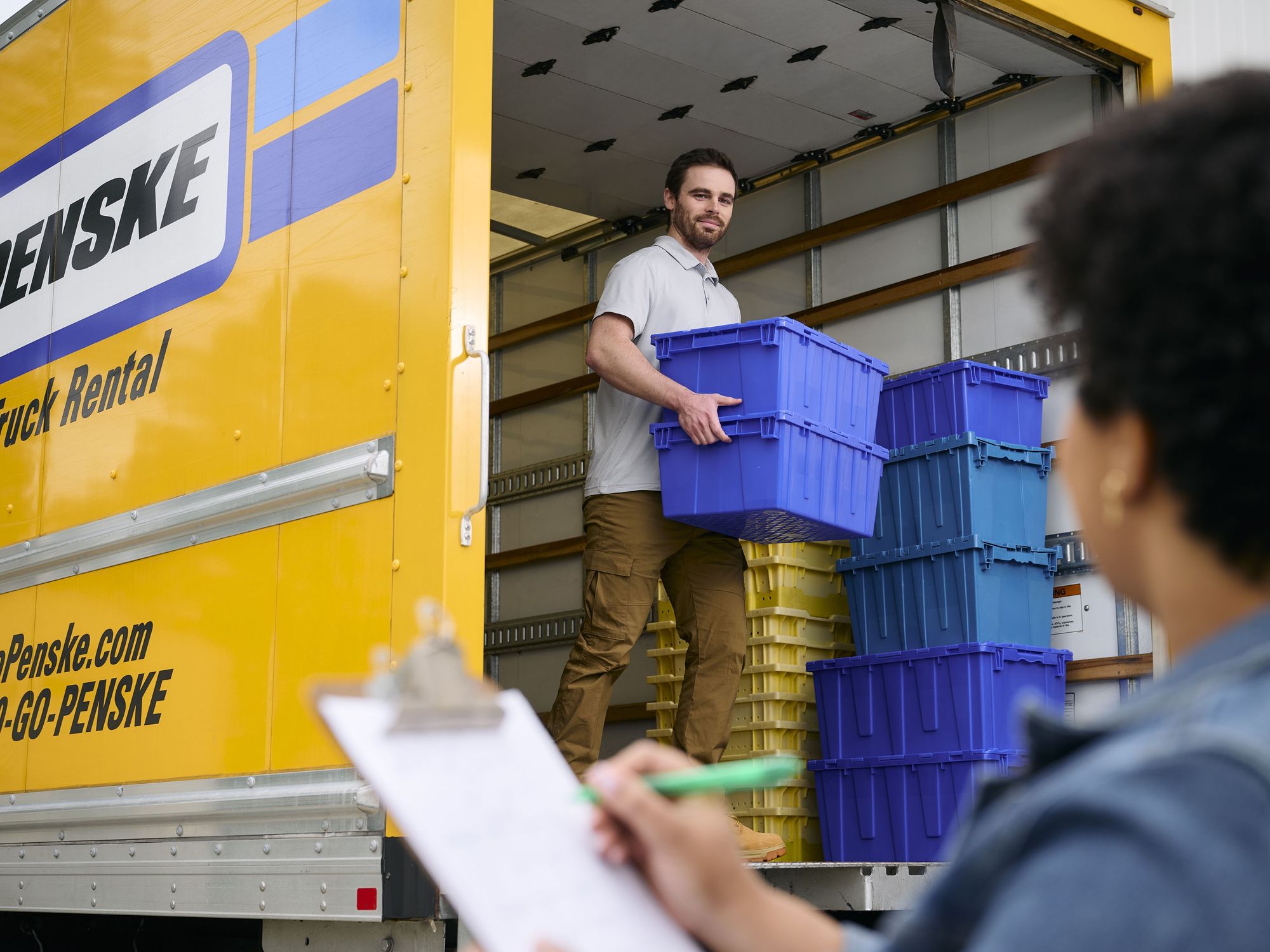 A man unloads crates from a yellow Penske truck while a woman marks things off on a checklist.