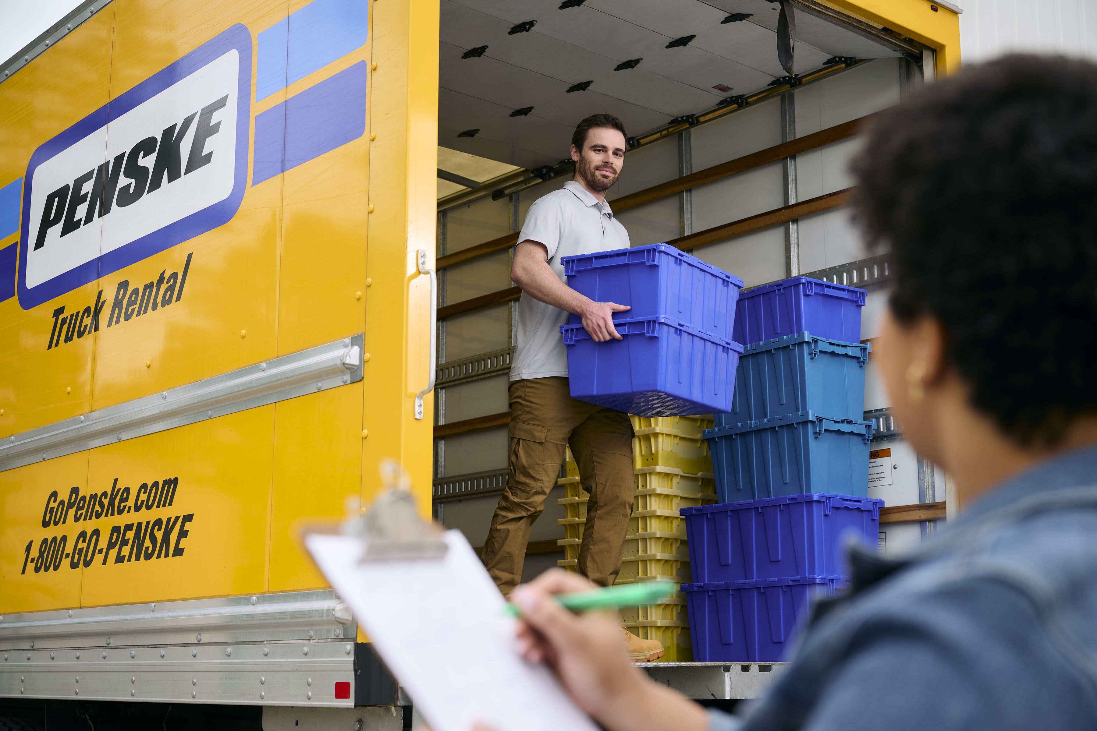 A man unloads crates from a yellow Penske truck while a woman marks things off on a checklist.