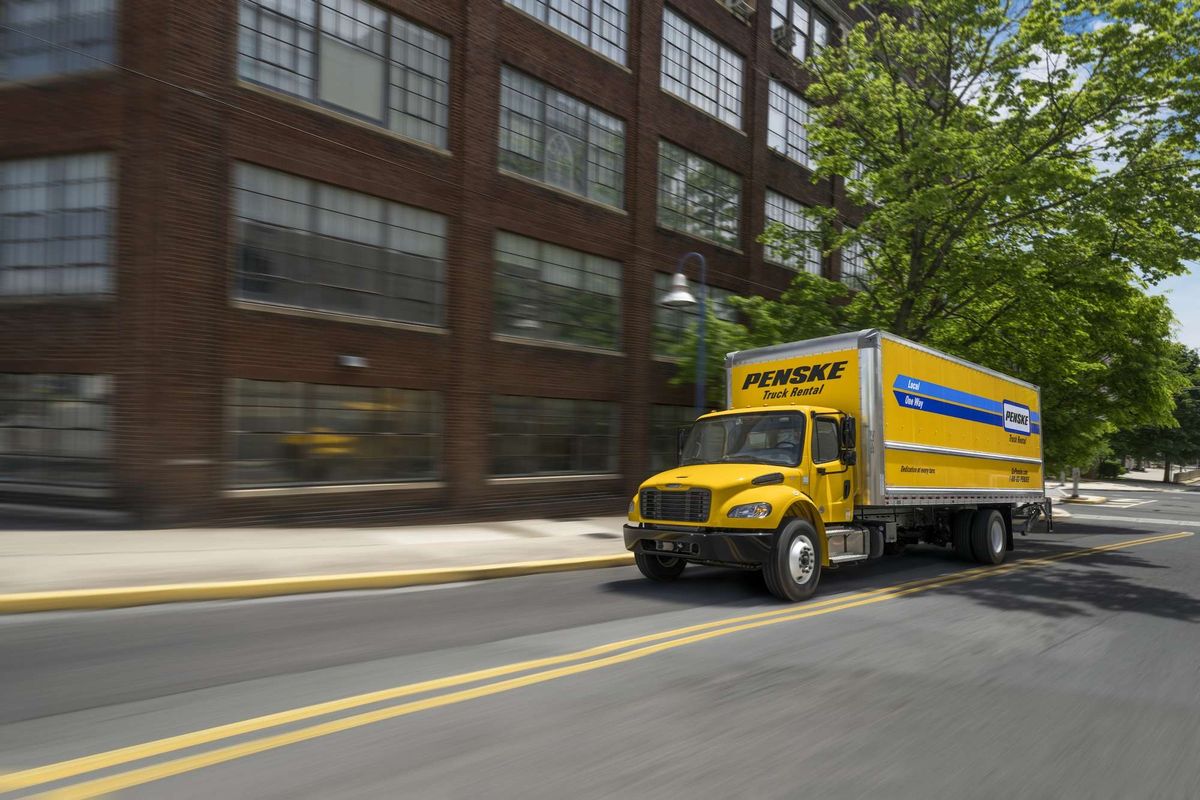 A medium-duty yellow Penske truck driving on the road next to a brick building.