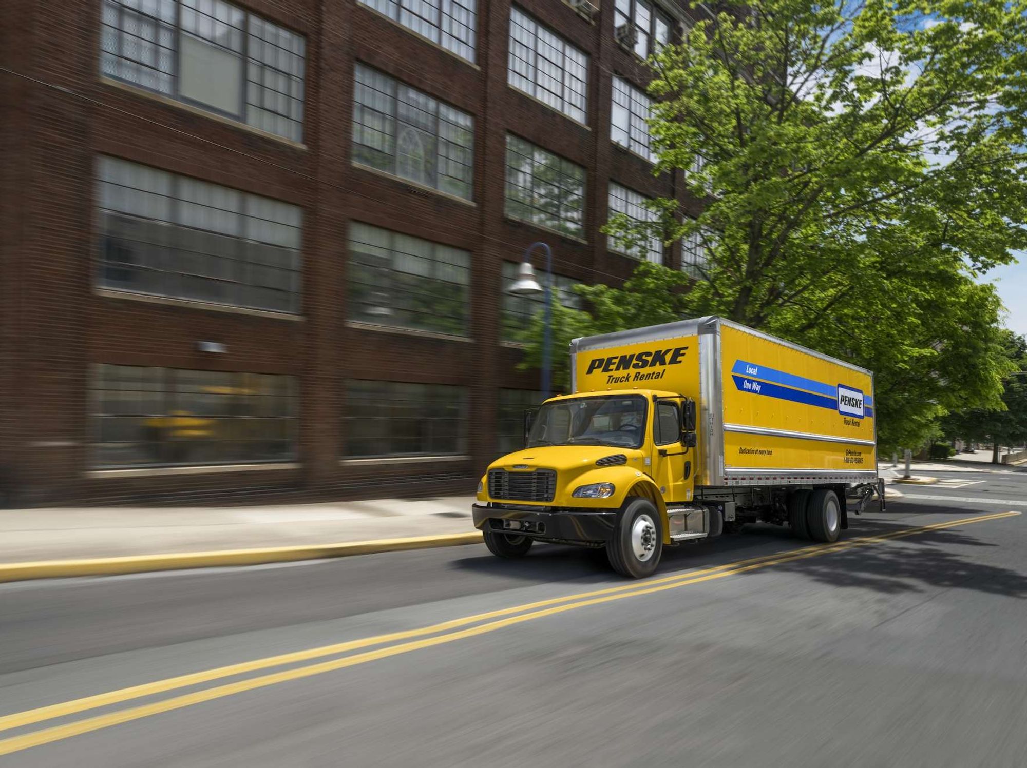 A medium-duty yellow Penske truck driving on the road next to a brick building.