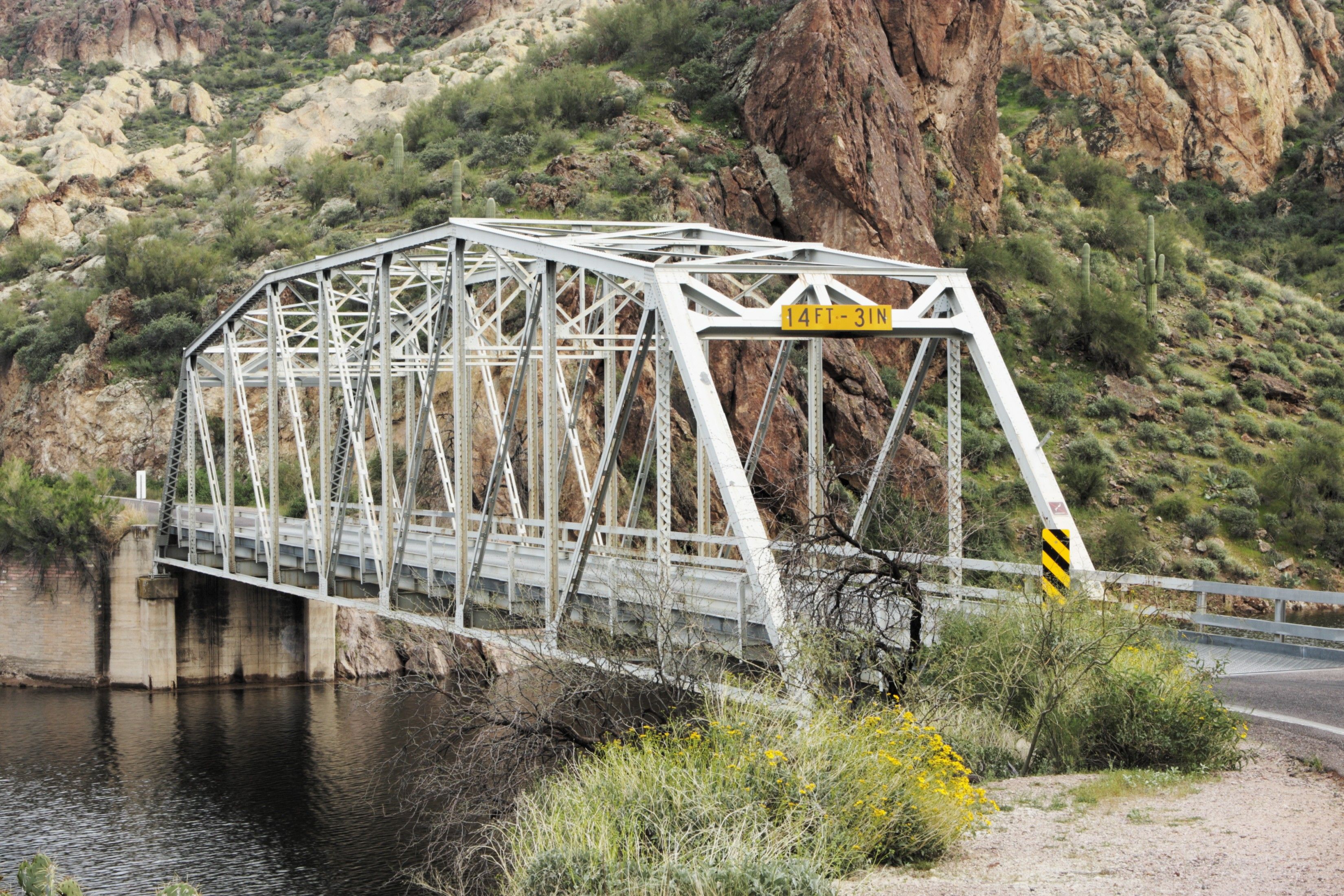 A metal bridge with a 14 ft - 3 in sign on it going over a river next to mountains.
