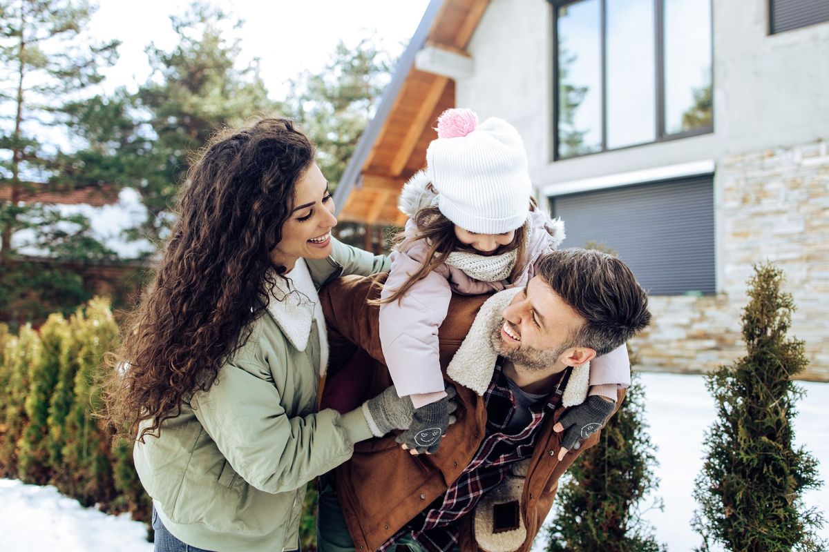 A mom, dad, and daughter have fun in the snow outside an a-frame house.