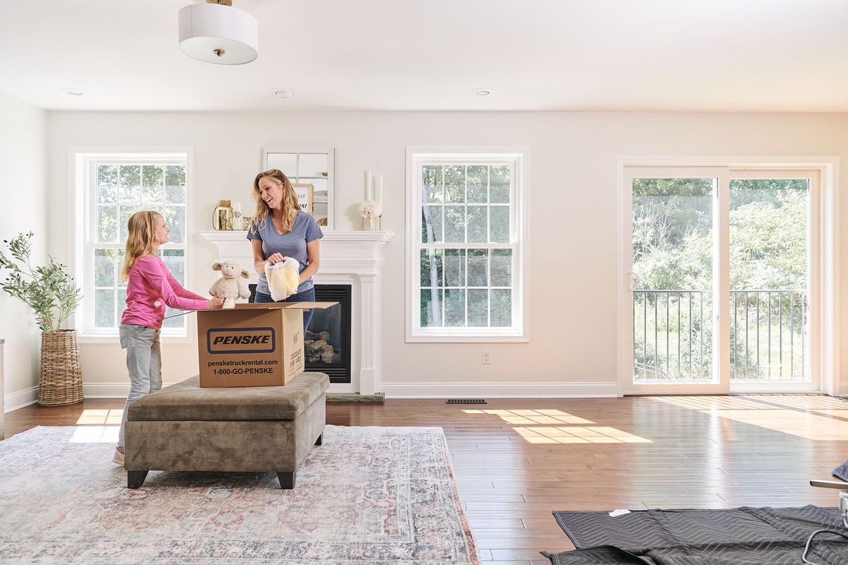 A mother and daughter stand in a nearly empty living room packing a Penske moving box.