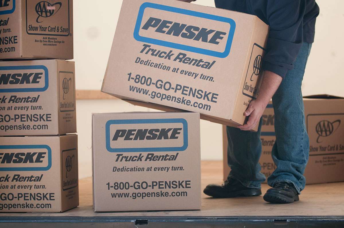 A person in blue jeans and a blue shirt bends over to lift a cardboard Penske box.