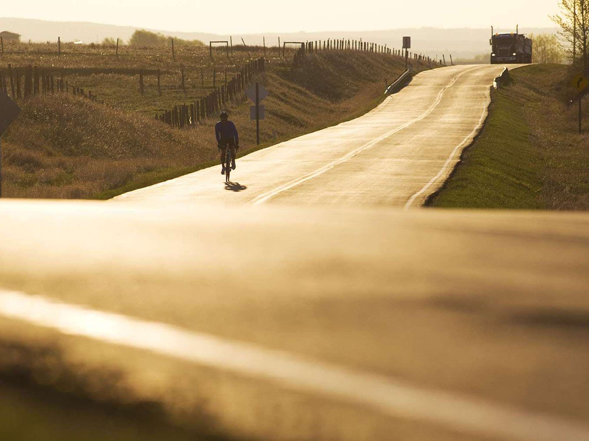 A person rides a bike down a road at sunset with a semi-truck behind.