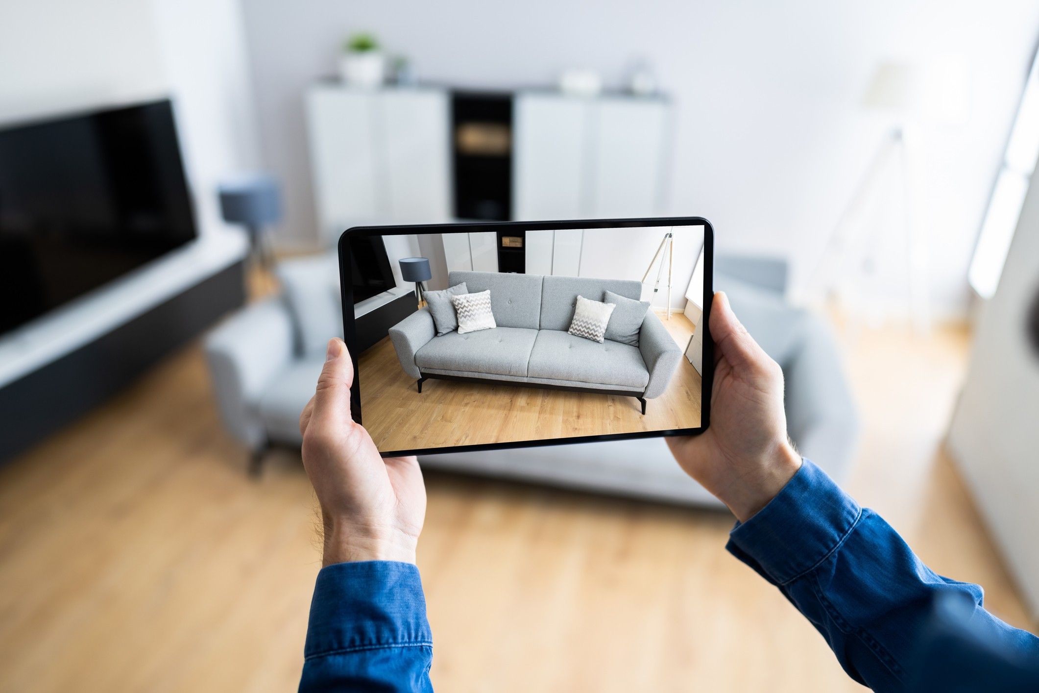 A person wearing a blue shirt takes a photo of a grey couch with a tablet device.