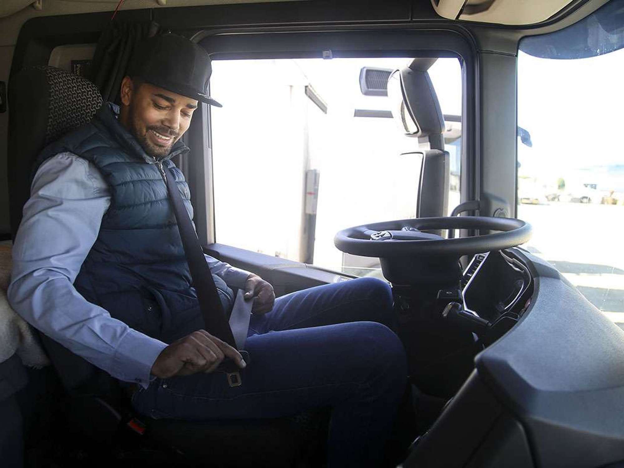 A truck driver in a blue vest and hat buckles his seatbelt while sitting in the cab of a semi-truck.