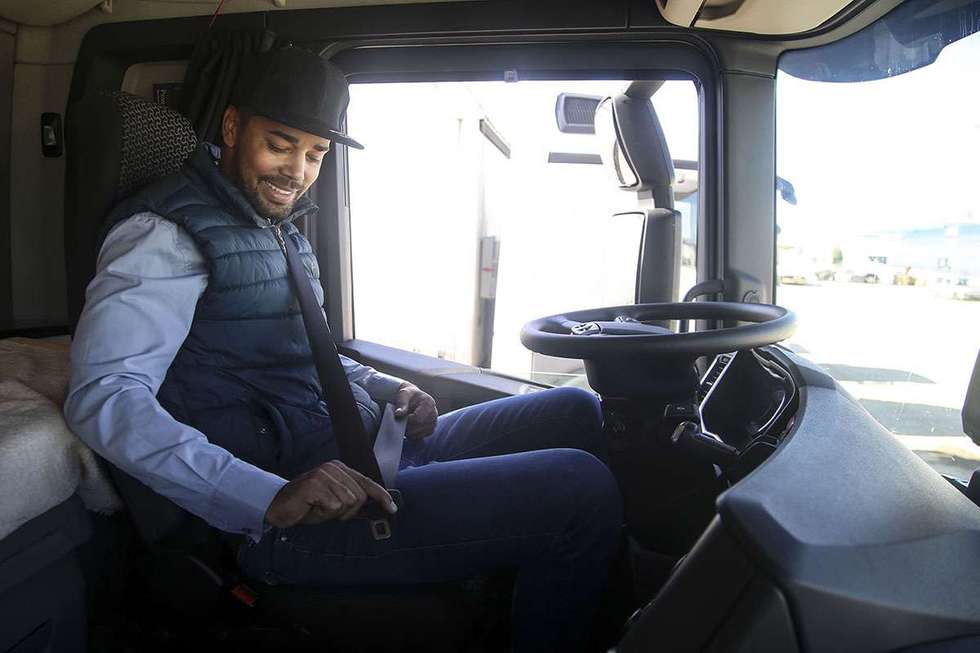 A truck driver in a blue vest and hat buckles his seatbelt while sitting in the cab of a semi-truck.