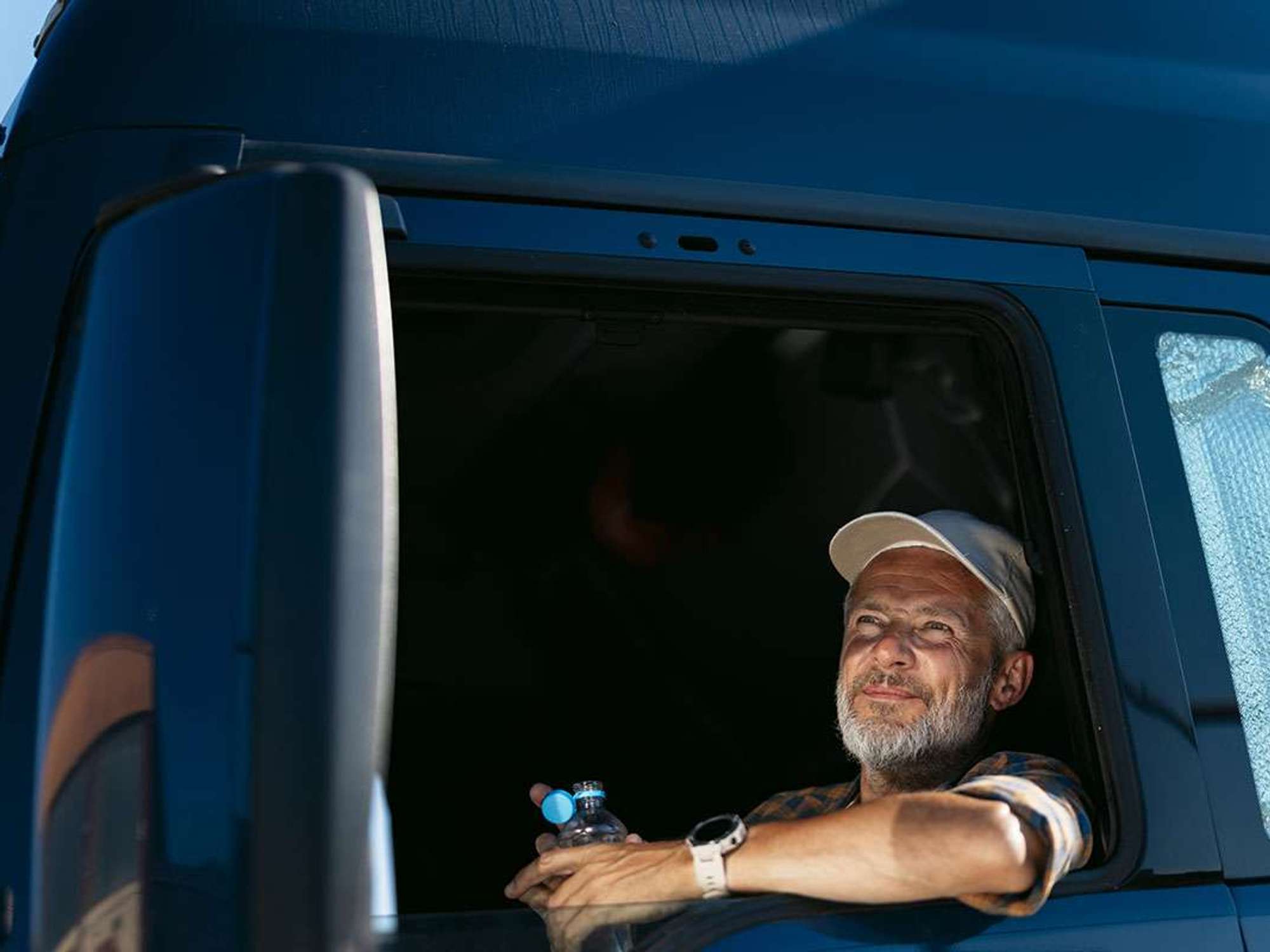 A truck driver in a truck cab window holding an open bottle of water.