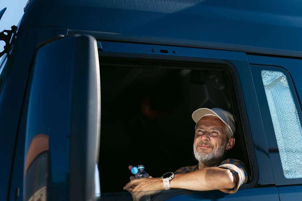 A truck driver in a truck cab window holding an open bottle of water.