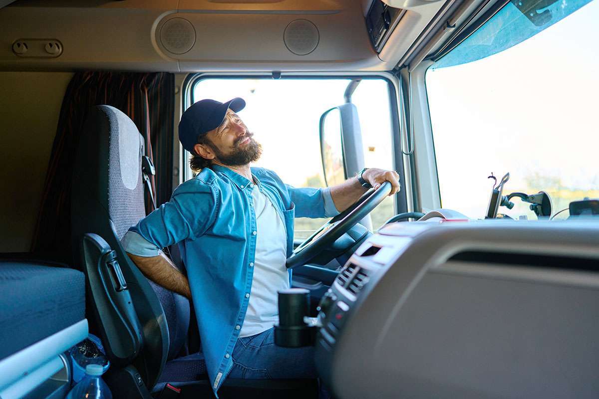 A truck driver sits in the cab of a semi-truck arching his back in pain.