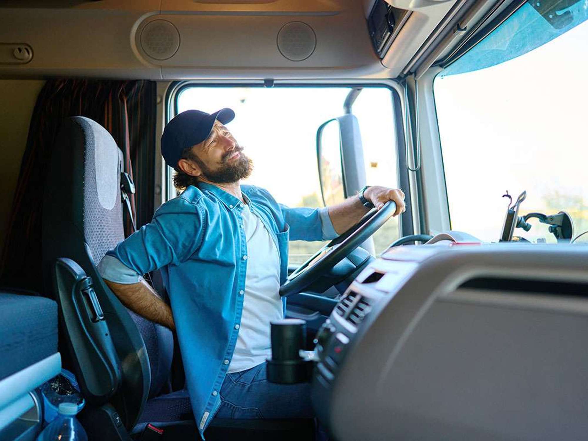 A truck driver sits in the cab of a semi-truck arching his back in pain.