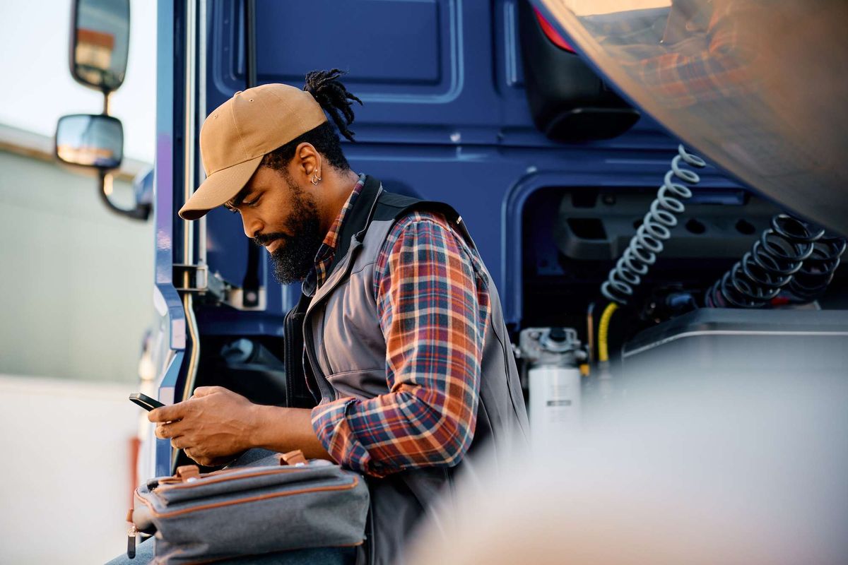 A truck driver sits on the side of his cab looking at a handheld device.