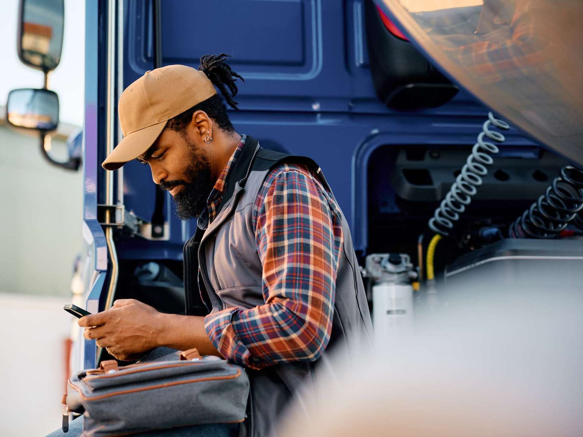 A truck driver sits on the side of his cab looking at a handheld device.