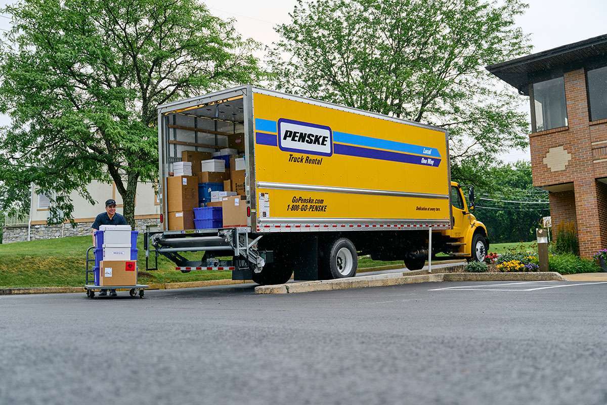 A truck driver unloads crates and boxes from the back of a parked yellow Penske box truck.