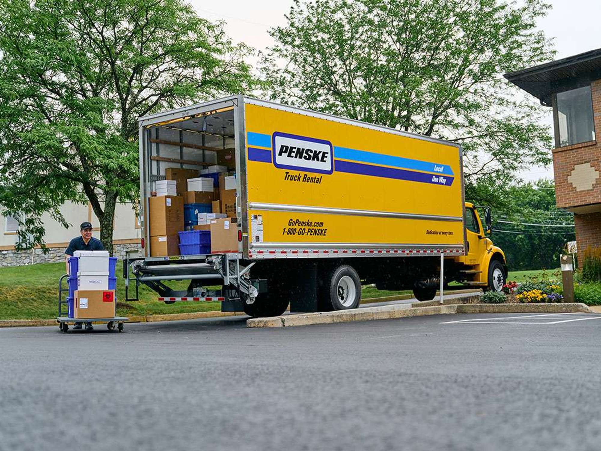 A truck driver unloads crates and boxes from the back of a parked yellow Penske box truck.