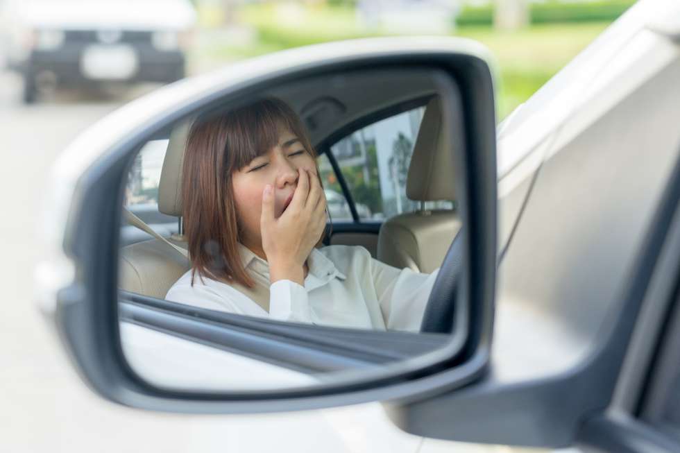 A vehicle side mirror showing a woman in a car yawning with her hand over her mouth.