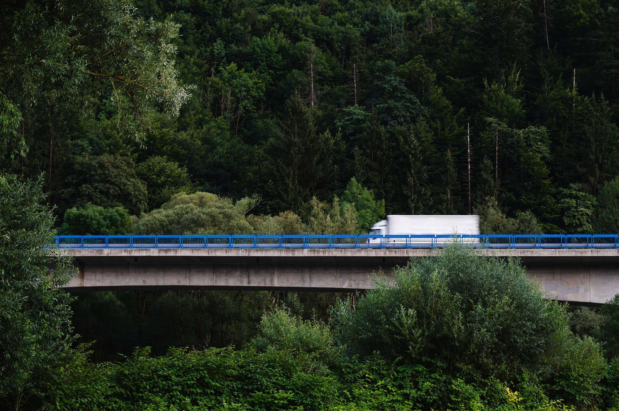 A white box truck drives across a bridge surrounded by a lush green forest.