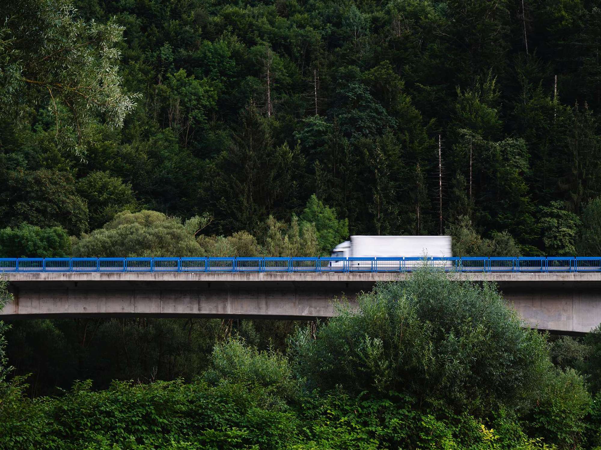 A white box truck drives across a bridge surrounded by a lush green forest.