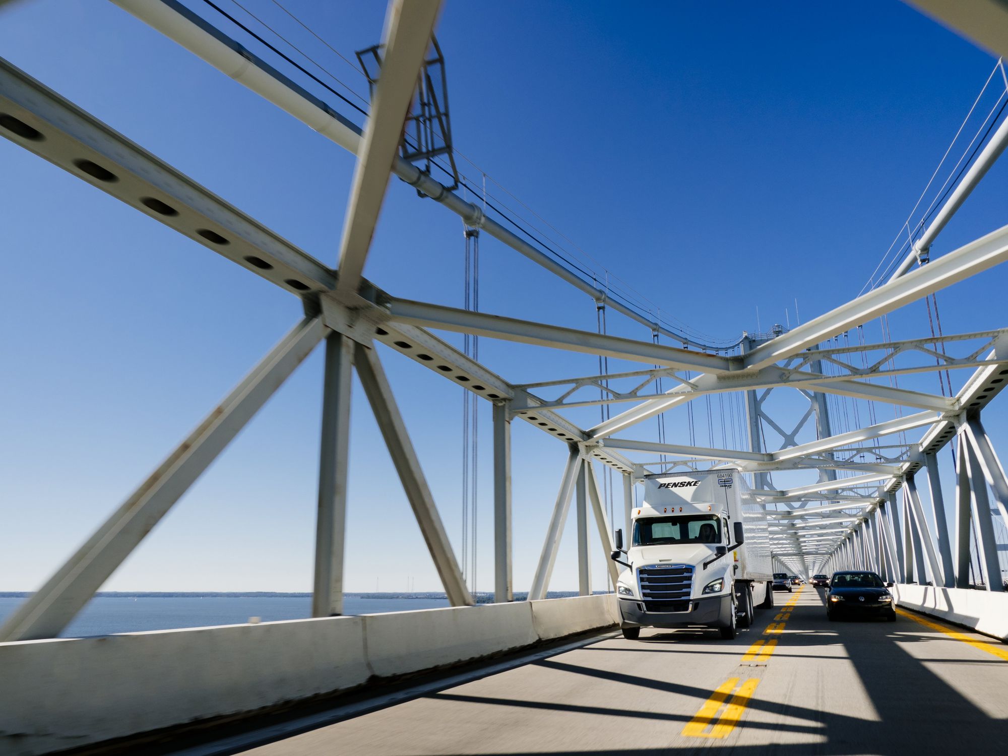 A white Penske semi-truck drives over a bridge on a sunny day.
