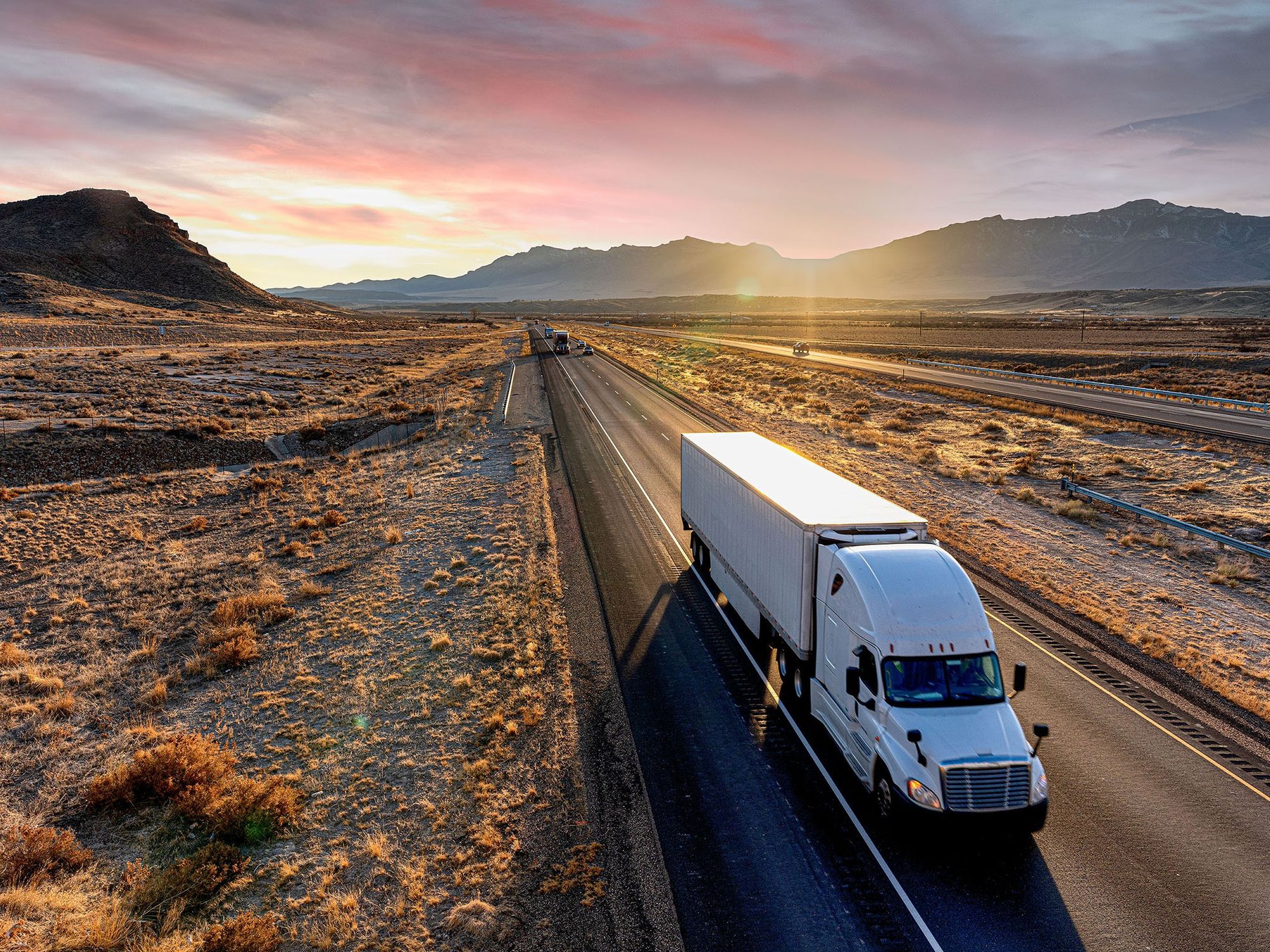 A white semi-truck drives down a desert road at dusk.