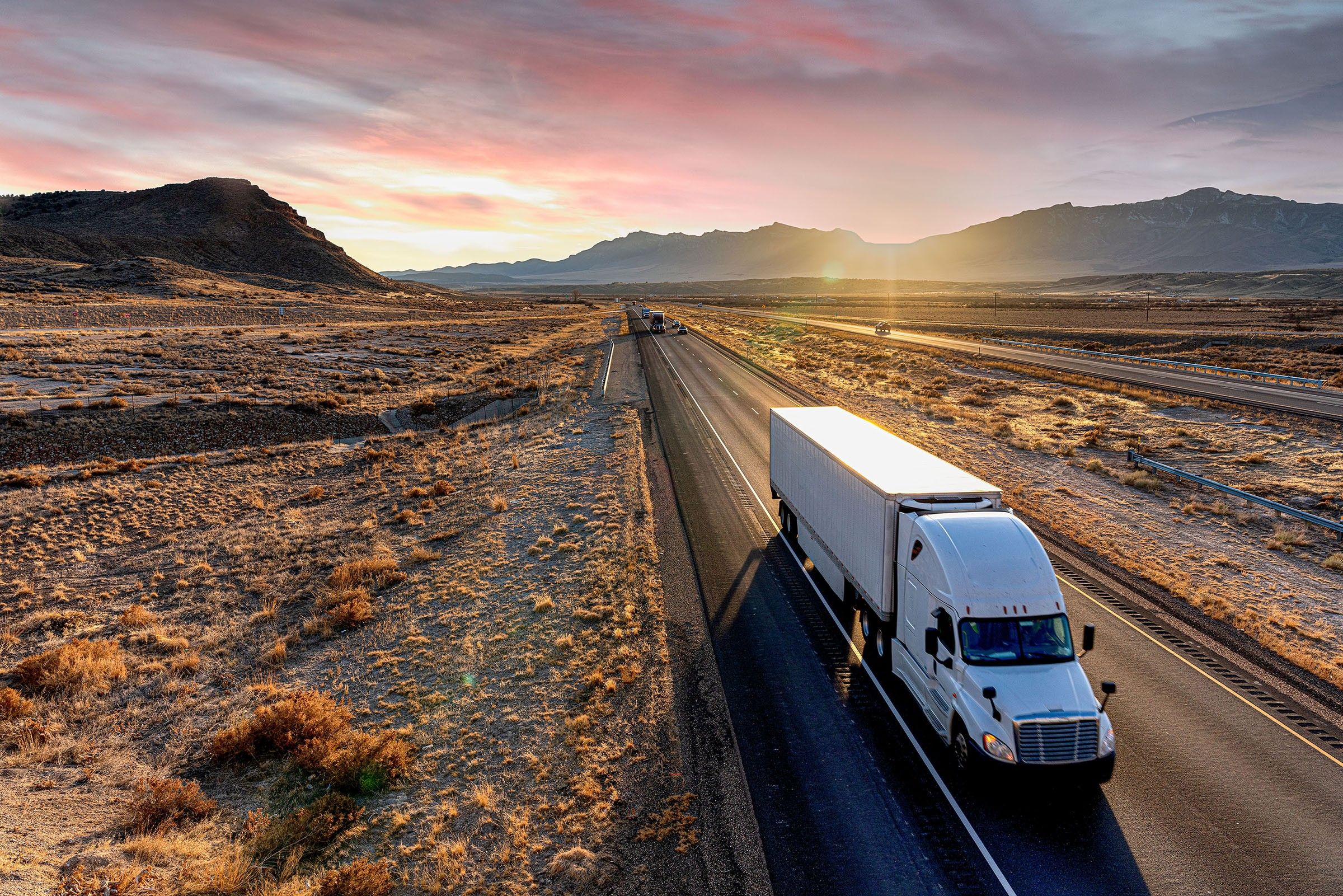 A white semi-truck drives down a desert road at dusk.