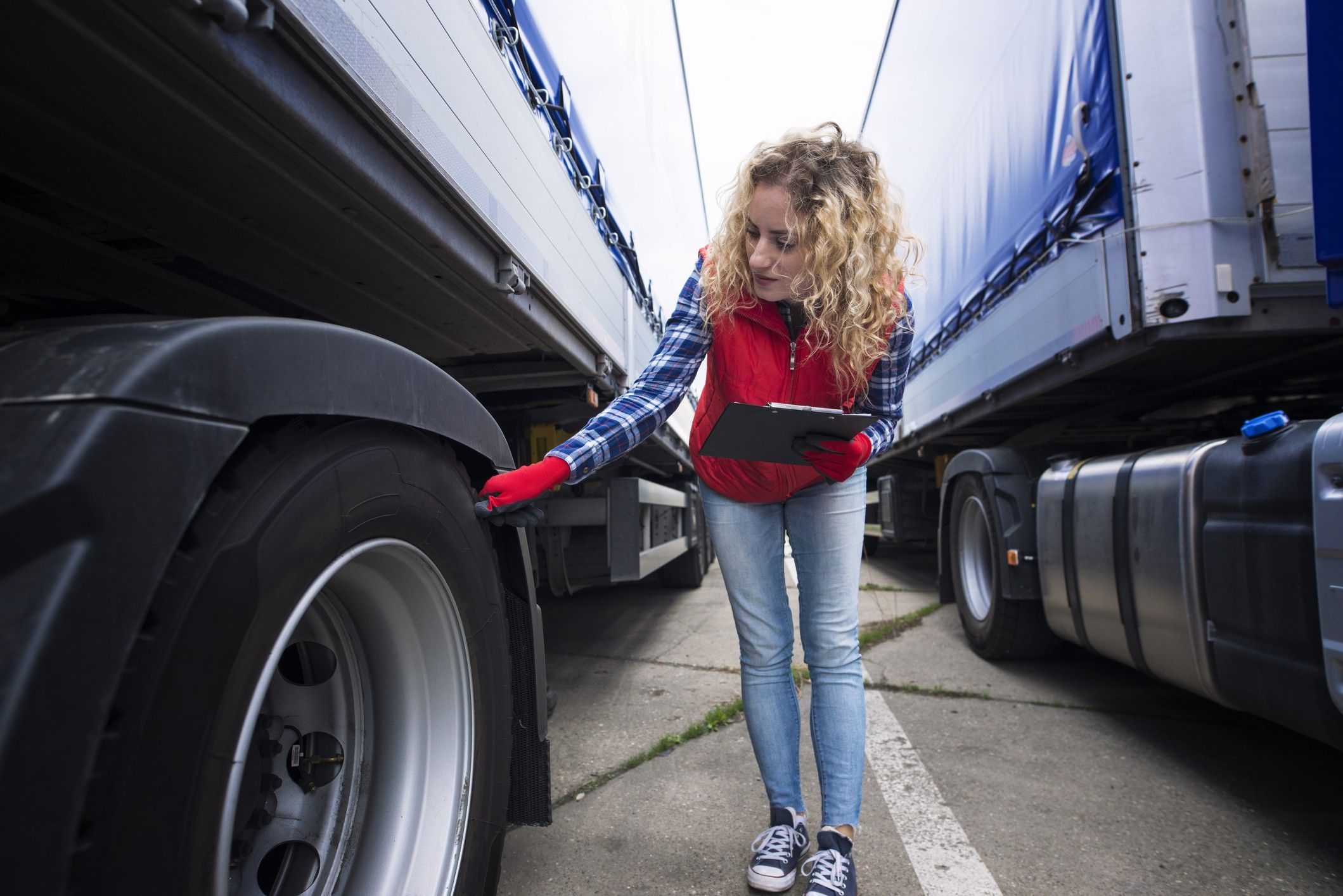 A woman holds a clipboard and checks the tire of a semi-truck.